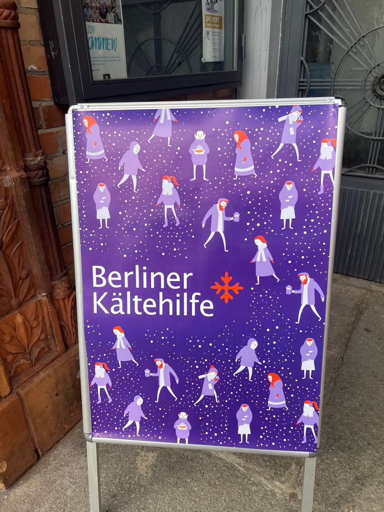 Ein Aufsteller der Berliner Kältehilfe mit dem Wimmelbild verschiedener Menschen im Winter steht vor dem Eingang in die Kirche.