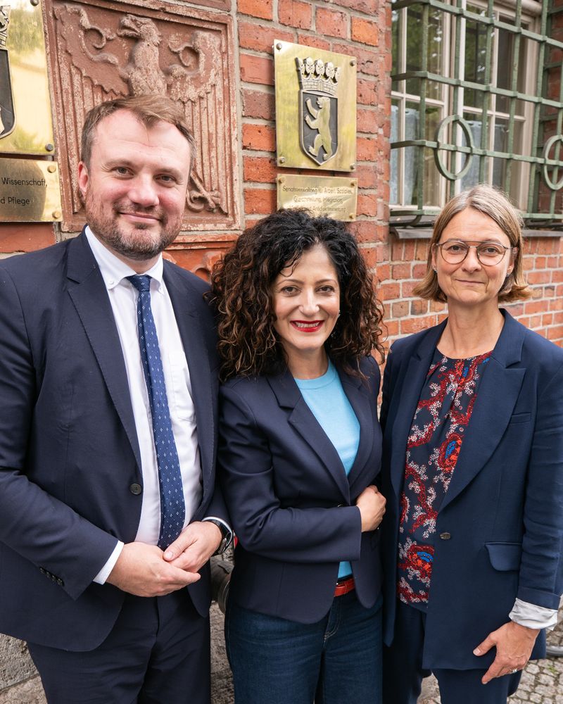 (v.l.n.r.) Minister für Wirtschaft, Arbeit, Energie und Klimaschutz von Brandenburg Daniel Keller, Senatorin für Arbeit, Soziales, Gleichstellung, Integration, Vielfalt und Antidiskriminierung Cansel Kiziltepe und Vorsitzende der DGB Berlin-Brandenburg Katja Karger stehen gemeinsam vor dem Gebäude der Berliner Senatsverwaltung ASGIVA in der Oranienstr. 106. Im Hintergrund sind ein Schild der Senatsverwaltung mit dem Wappen von Berlin und ein Adler an der Backsteinwand, der an den Wappen von Brandenburg erinnert, sichtbar.