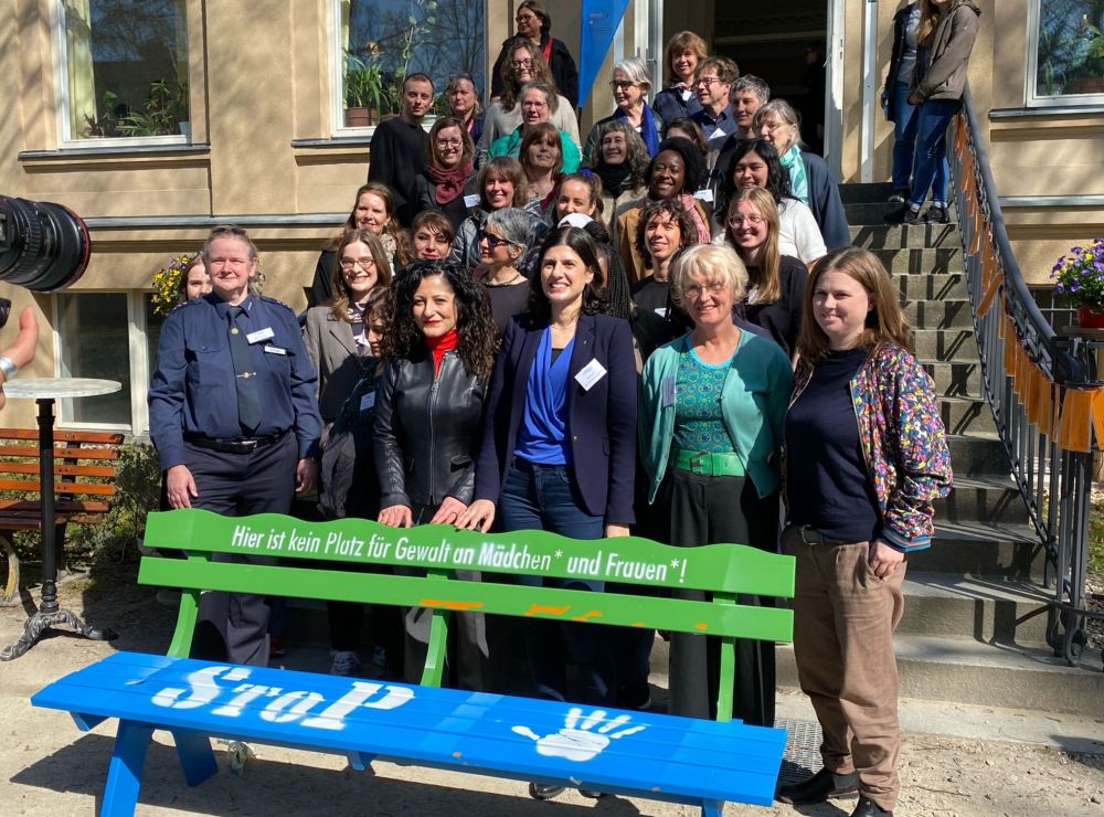 Ein Gruppenbild: Senatorin Cansel Kiziltepe, MdA Derya Çağlar, Bürgermeisterin Clara Herrmann, Prof. Dr. Sabine Stövesand und andere Teilnehmende des Fachtags stehen im Hinterhof des „Nachbarschaftshaus Urbanstraße“ hinter einer blau-grünen Sitzbank mit dem aufgesprühten Wort „StoP“ und dem Bild einer Handfläche sowie dem Text „Hier ist kein Platz für Gewalt an Mädchen* und Frauen*!“