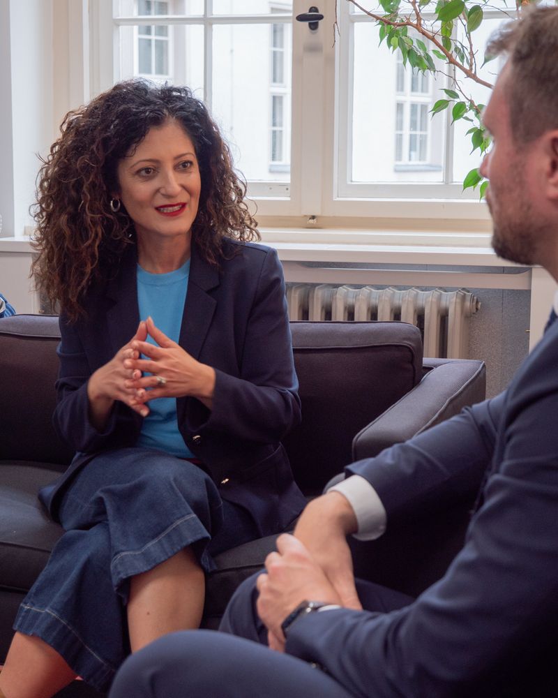 Senatorin Cansel Kiziltepe spricht gestikulierend mit dem Minister Daniel Keller, der nur teilweise im Bild zu sehen ist. Die Szene spielt sich im Amtszimmer der Senatorin mit großem Fenster und Pflanzen im Hintergrund ab.