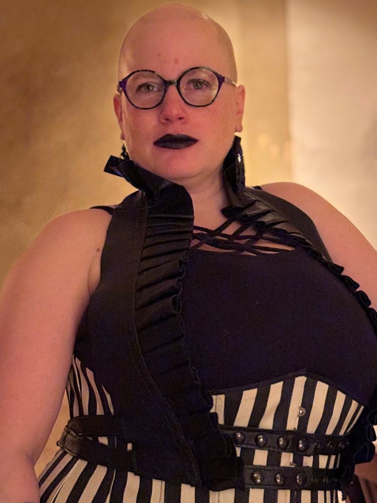 Jenn looking directly into the camera. She's wearing glasses, black lipstick, long dangly black earrings, a striped corset, and a black leather shrug/harness thing over top of the corset.