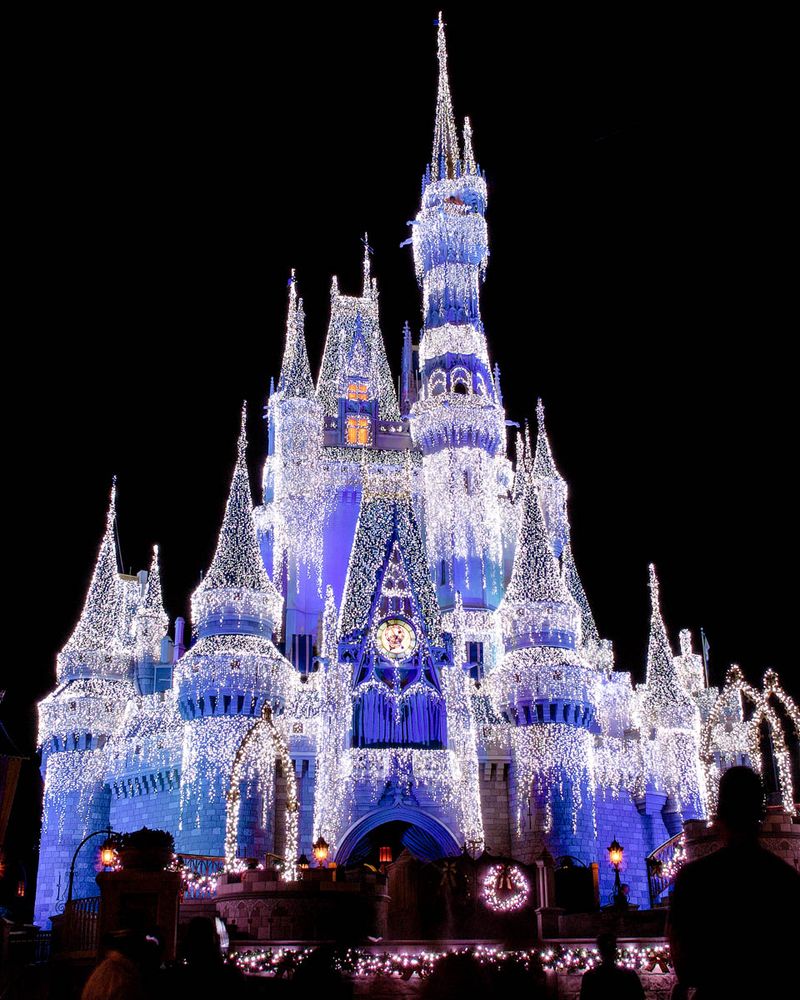 Cinderella's Castle at Magic Kingdom illuminated by thousands of shimmering white and blue lights during the holiday season. The castle is glowing against the night sky, with icicle-like lights draped elegantly over its towers, creating a magical and enchanting atmosphere. Silhouettes of visitors can be seen admiring the view.