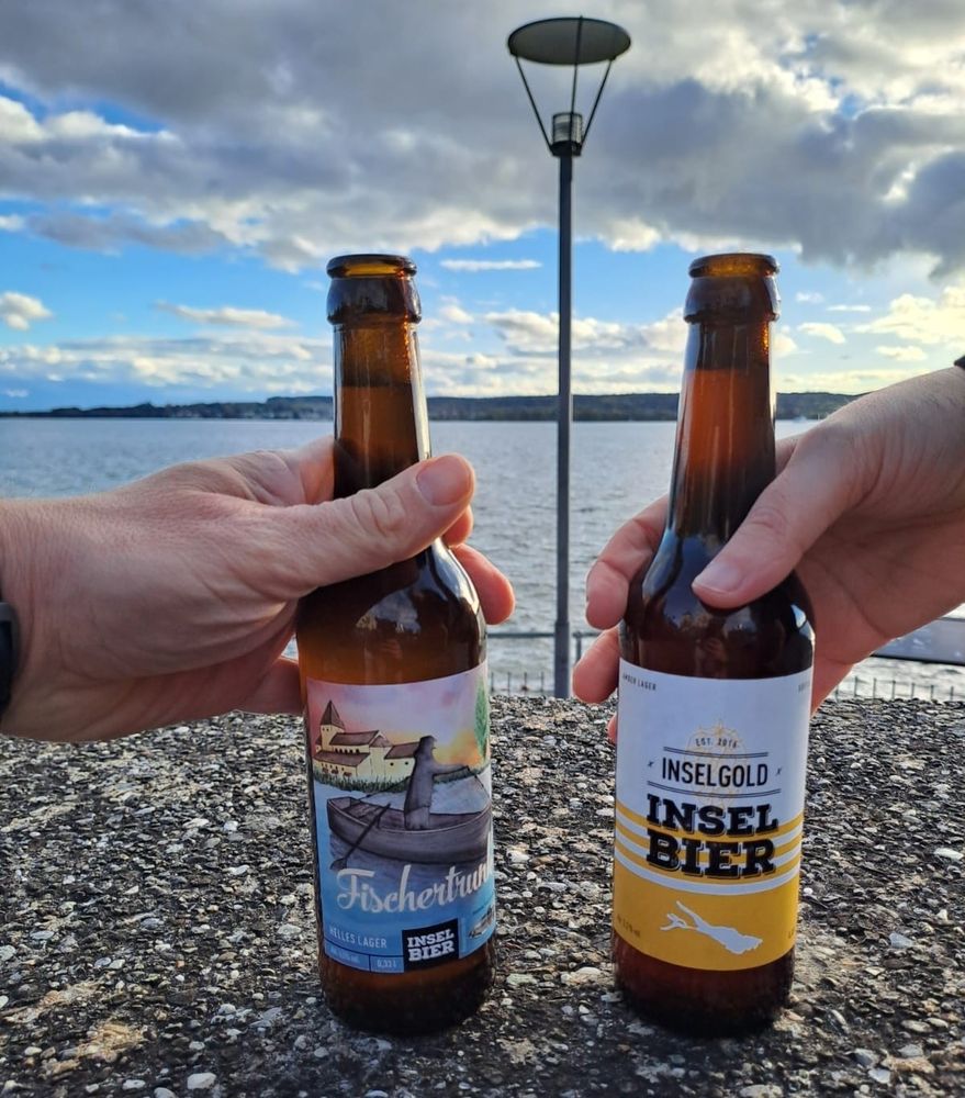 Selfie von Florian und mir. Wir lächeln in die Kamera und stoßen mit zwei Bierflaschen an. Hinter uns ist der Bodensee über dem gerade die Sonne untergeht.