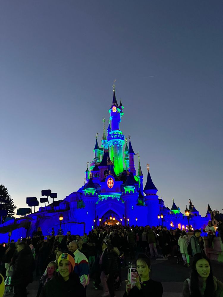 Het is donker buiten, je ziet het kasteel in Disneyland Parijs, dat blauw met groen verlicht is. Op de voorgrond staan een hoop mensen in de ruimte voor het kasteel. Naast het kasteel zijn bomen te zien.