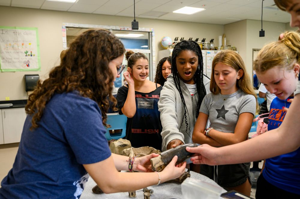 a group of what look like middle school girls are standing at a table to examine a large fossil held by an adult in what looks like a classroom