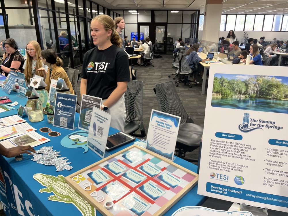 One person stands and several sit behind a table in a public space that displays many small information signs and cards while students are sitting at work stations in the background in what looks like a library
