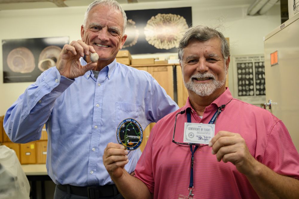 two older men are holding fossils and cards of some kind that are slightly blurry to read while standing for a portrait in a scientific fossil lab of some kind