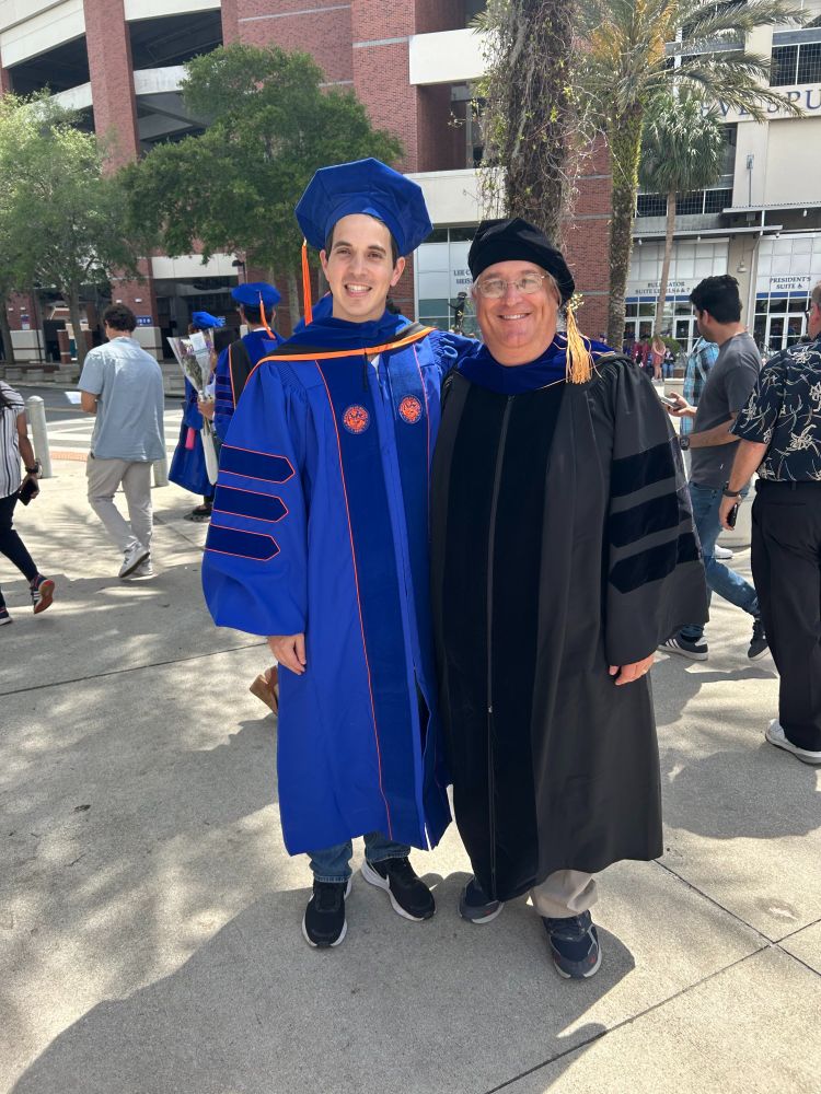 two people are standing outside wearing traditional university graduation robes, one in black and the other in dark blue