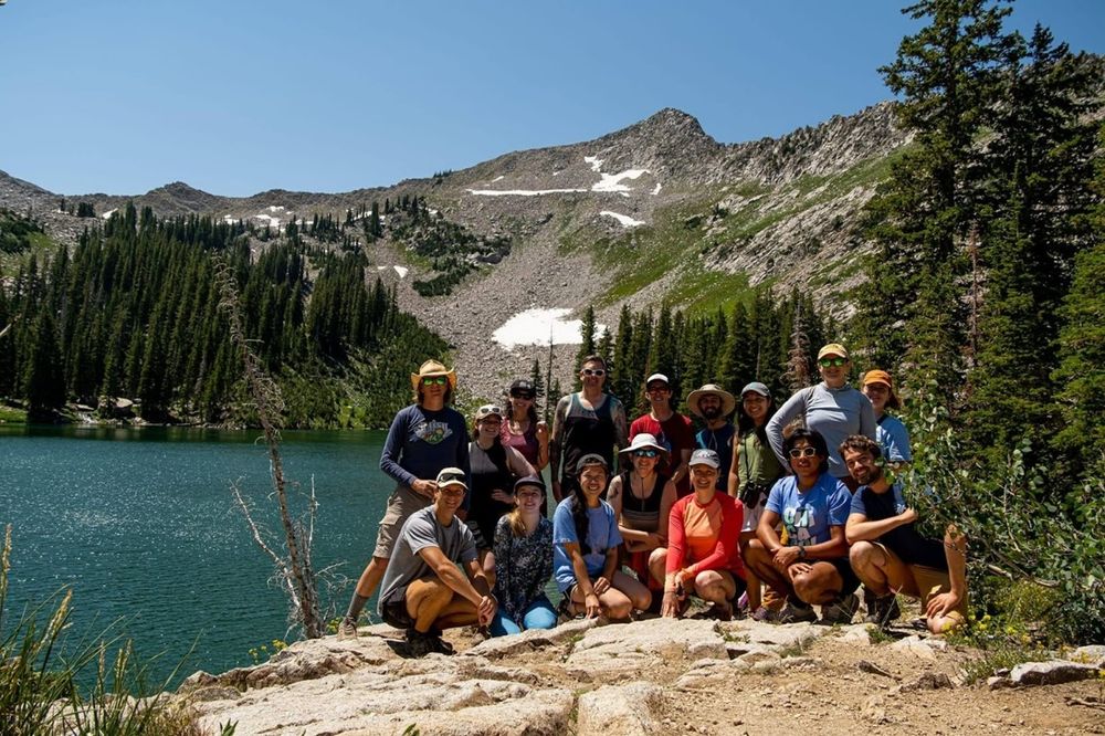 a group of over a dozen people in outdoors clothing are posing together on a rocky crest with a lake and rocky mountain behind them against a clear blue sky