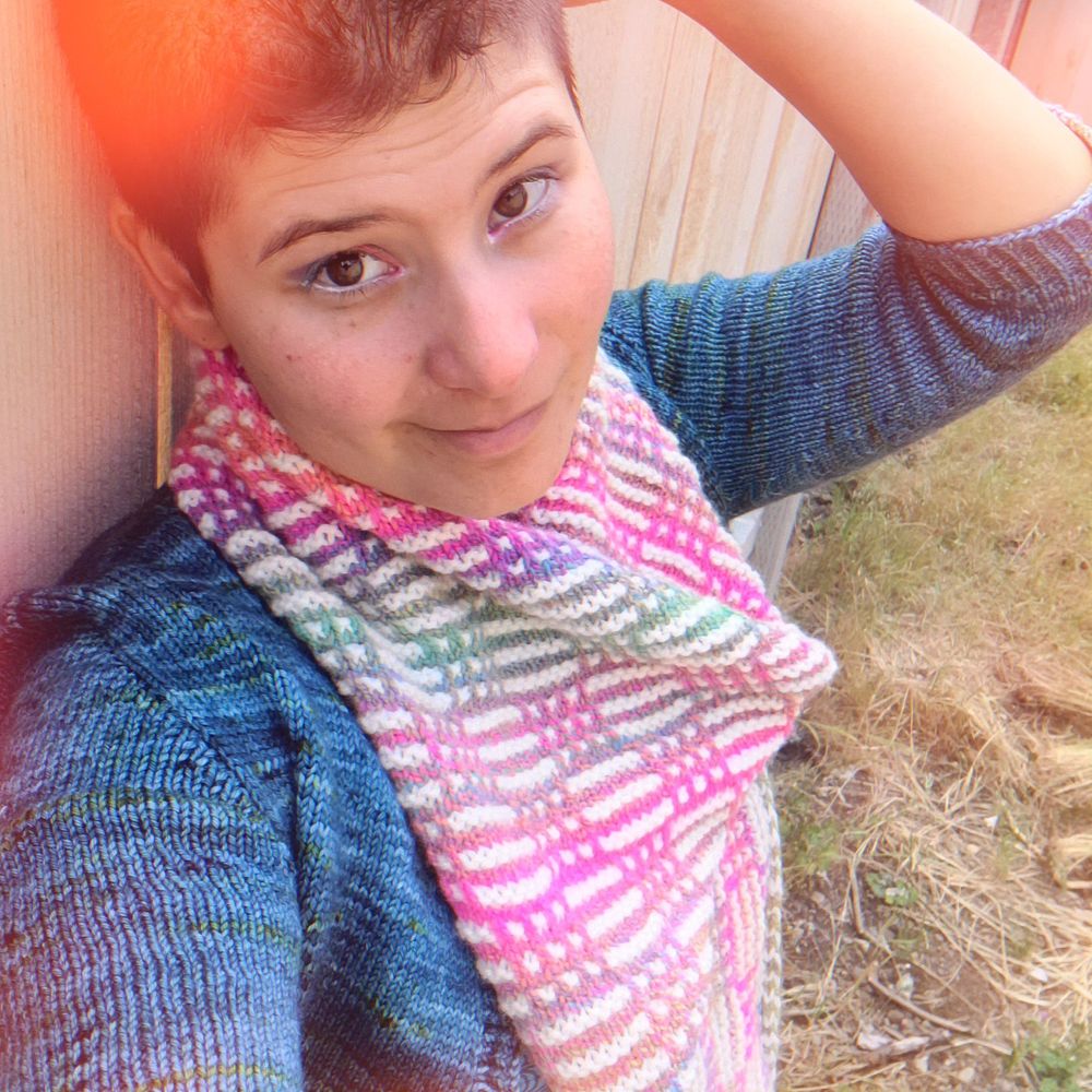 A mohawked femme/afab person poses  wearing a rainbow, mosaic/slip stitch knit cowl