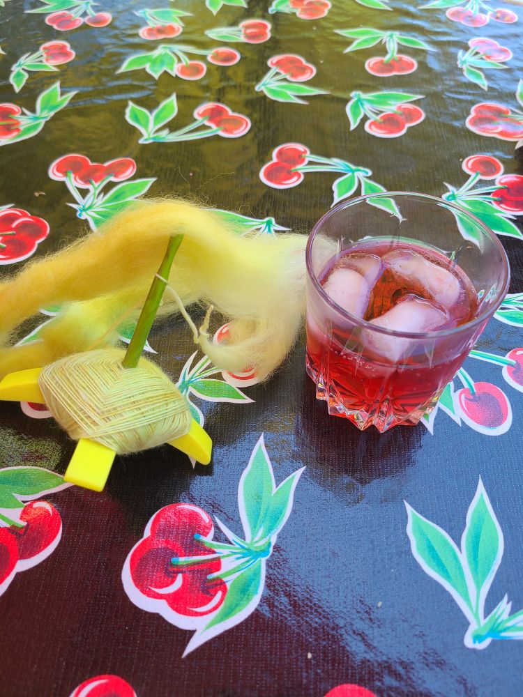 A green Turkish spindle with green fiber and singles sits on an oilcloth tablecloth next to a tumbler of jamaica