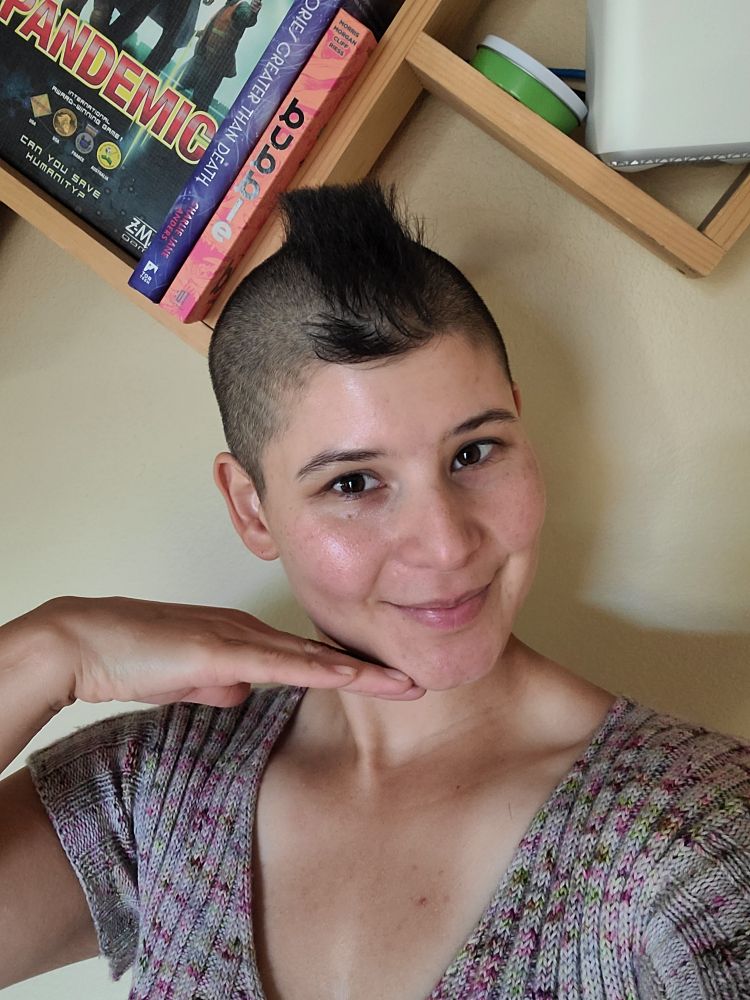 A Latina woman with a very cropped mohawk wearing a speckled handknit Ripple Crop Top in gray yarn stands in front of a bookshelf