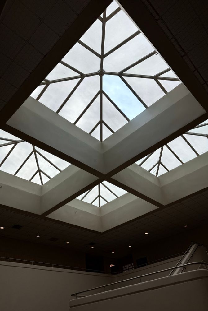 Vista hacia arriba del tragaluz geométrico dentro del Tribunal de Humacao en Puerto Rico. El techo muestra cuatro estructuras de vidrio en forma de pirámide, organizadas en cuadrado, con paneles triangulares y un centro metálico. El cielo está nublado con un leve tono azul. Las paredes blancas y la iluminación empotrada crean un ambiente minimalista e institucional.
————
View looking up at a geometric skylight inside the Humacao Tribunal in Puerto Rico. The ceiling features four pyramid-shaped glass structures arranged in a square, each with triangular panes and a central metal hub. The sky above is overcast with hints of blue. The white walls and recessed lighting below add to the minimalist, institutional atmosphere.