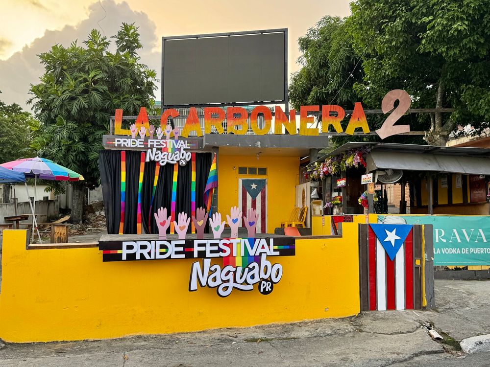 A bright yellow building named “La Carbonera 2” is decorated for the Pride Festival in Naguabo, Puerto Rico. A small stage with black curtains and rainbow accents displays the event name alongside colorful raised hands. A door and gate are painted with the Puerto Rican flag, and a rainbow flag hangs beside the entrance. A food kiosk with flowers and pride signs is attached to the right. The sky is overcast, and a colorful umbrella shades an outdoor area with picnic tables.

Un edificio amarillo brillante llamado “La Carbonera 2” está decorado para el Pride Festival en Naguabo, Puerto Rico. Un pequeño escenario con cortinas negras y acentos arcoíris muestra el nombre del evento junto a manos coloridas levantadas. Una puerta y un portón están pintados con la bandera de Puerto Rico, y una bandera del orgullo cuelga al lado de la entrada. Un kiosco de comida con flores y letreros de orgullo está adjunto a la derecha. El cielo está nublado, y una sombrilla multicolor cubre una zona de mesas al aire libre.