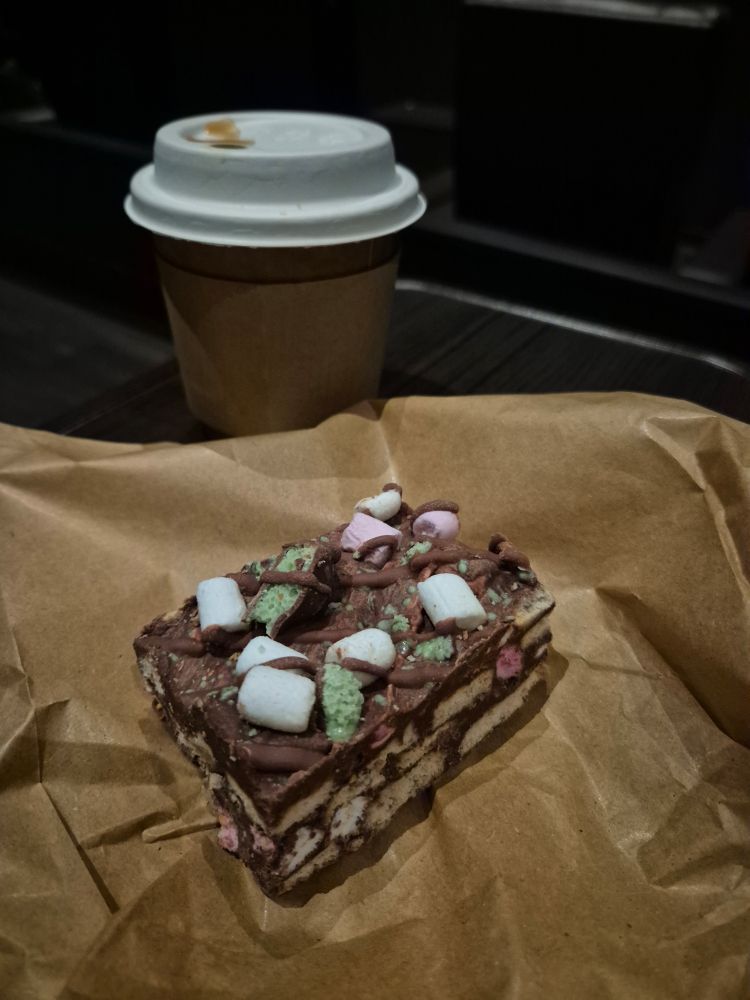 A takeaway cup and a slice of rocky road on a brown paper bag in a dimly lit cinema 