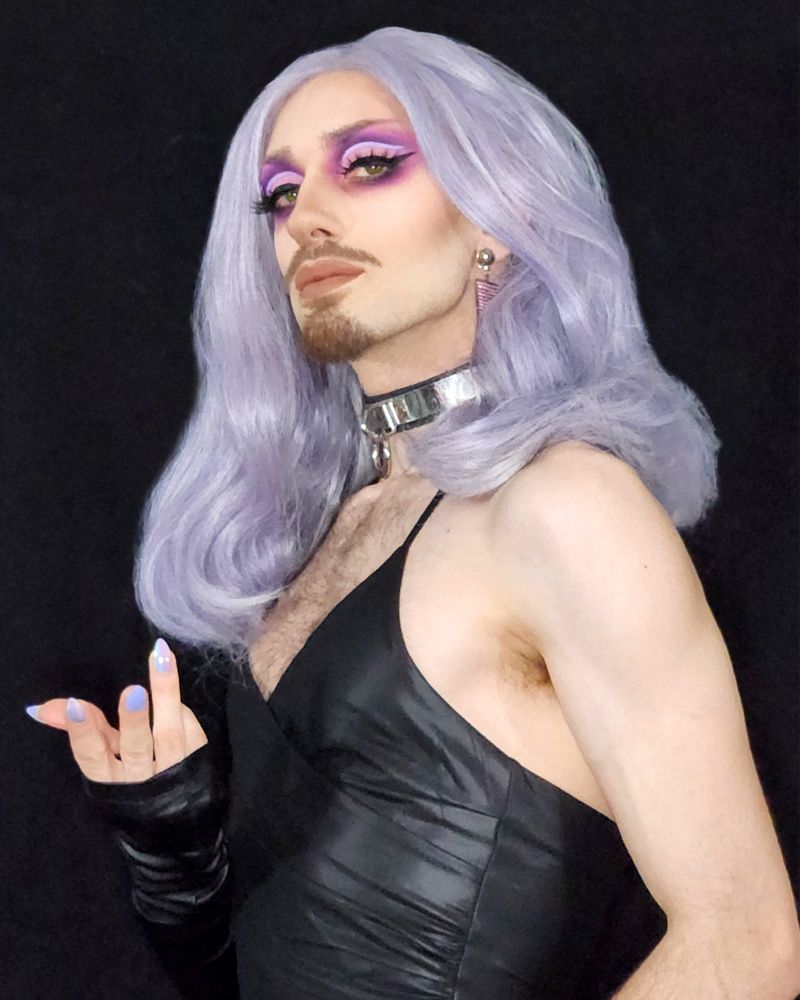 Duchess, a bearded drag artist, beckoning you in three-quarter profile wearing a black ruched vinyl spaghetti-strap mini dress, matching fingerless opera glove, silver metal choker, and pink rhinestone covered triangle earrings. Makeup features a vibrant purple and pink cut crease floating above spiked lashes along a large eyeliner wing and a neutrally nude truffle lip. Ice lavender hair drapes in large waves just past their shoulders. I look very pretty I promise.
