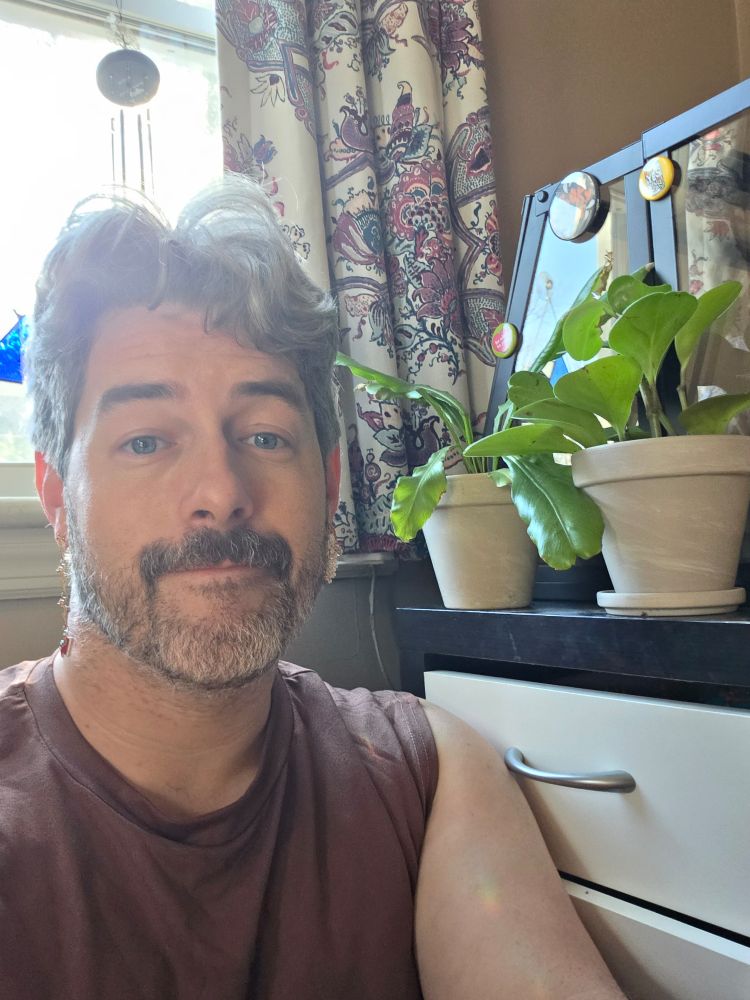 Me, a man with a greying beard and perpetual bedhead, sitting next to a small dresser with indoor plants and decorative magnets