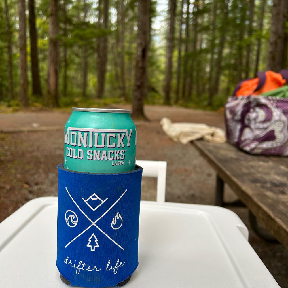 A can of Montucky Cold Snacks beer in a Drifter Life koozie sits on top of a cooler at a campground in the north Cascades National Park