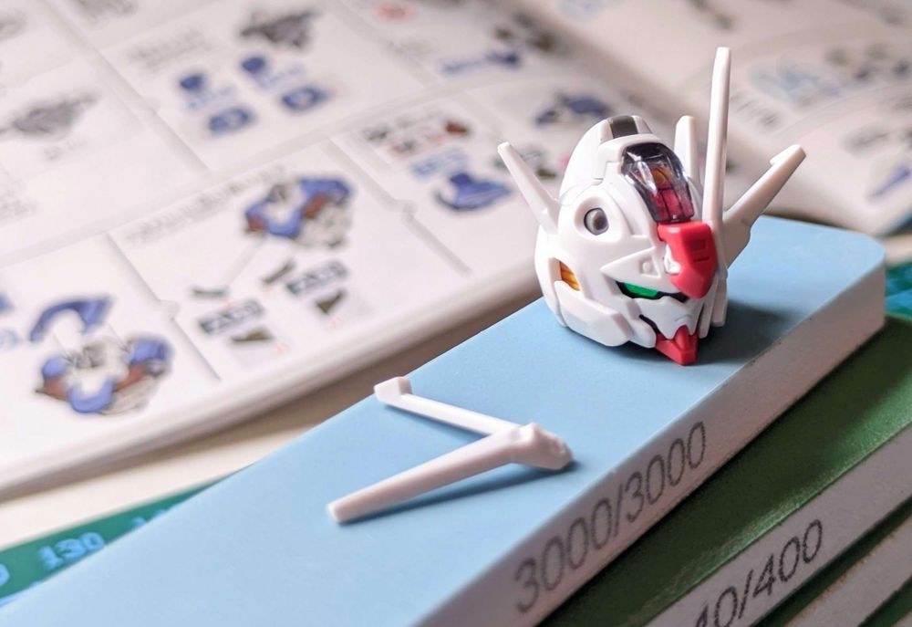 A photo of a gunpla head with a broken V-fin, sitting atop a pile of sandpaper sticks.