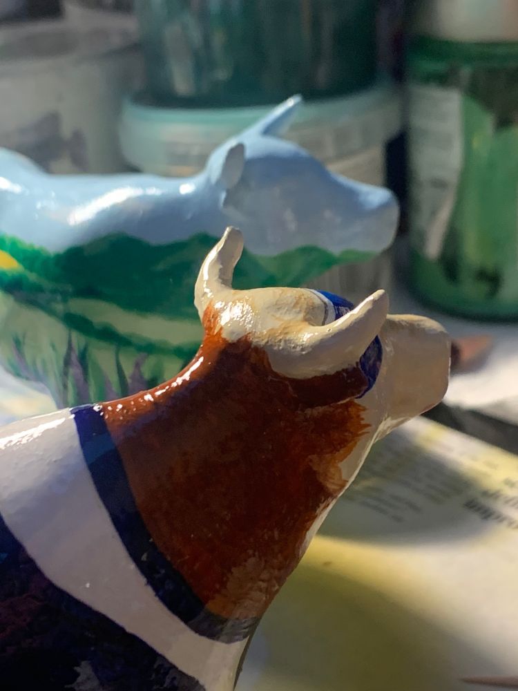 Sandra Russell art - close up of two ox figurines painted by Sandra - the closest has blue sunglasses and wears a white and dark blue top. The one in the background is a landscape with blue sky and green fields.