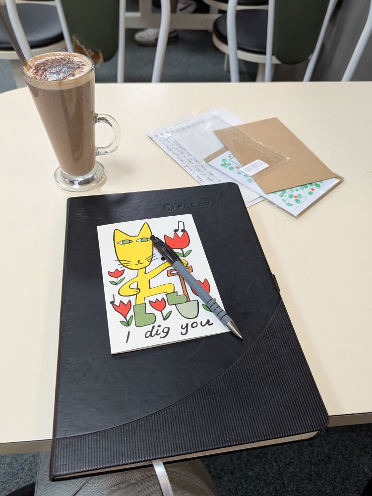 A latte with cocoa powder sits on a white table next to a digital drawing tablet displaying a colorful illustration of a yellow cat with gardening tools and red tulips, with the text 'I dig you.' A stylus pen rests on the tablet, and envelopes are scattered nearby.