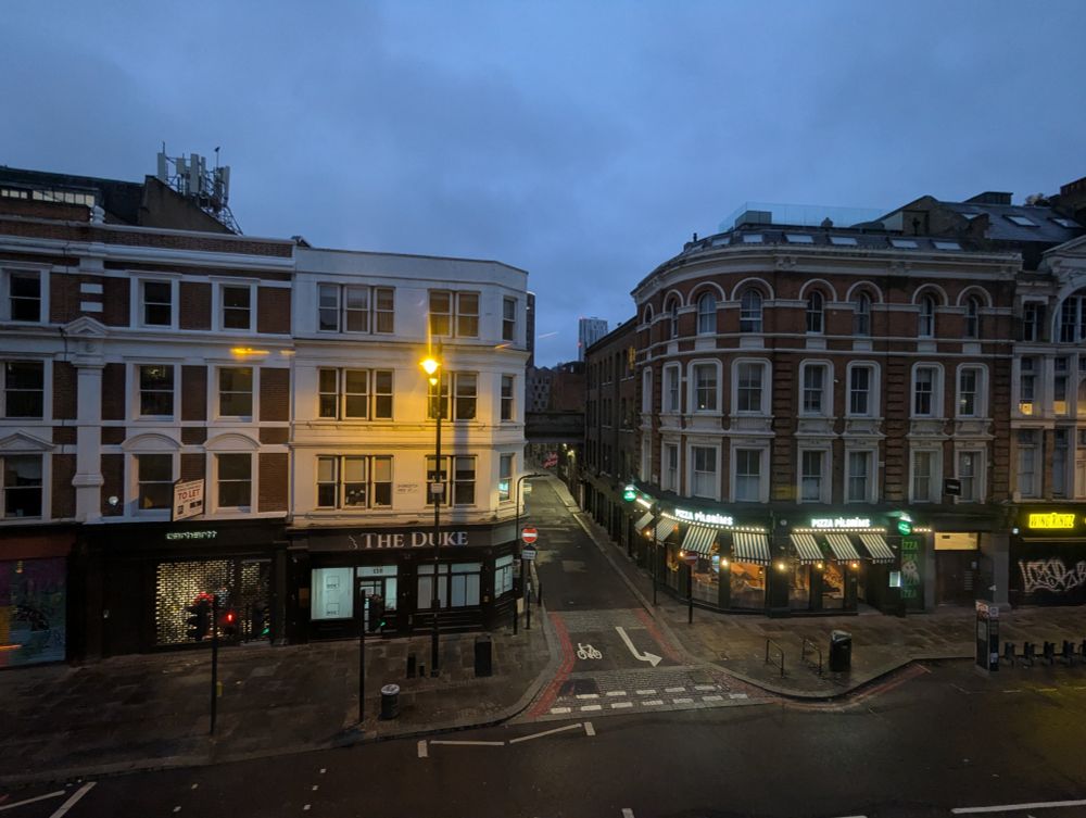 A quiet London street corner in Shoreditch at dawn, showing the intersection of two roads with characteristic Victorian and Edwardian buildings. On the left is a white-painted building with 'The Duke' pub at street level and a Carhartt store, while on the right stands a handsome curved red brick building with white stone detailing and arched windows, housing a Pizza Pilgrims restaurant with green and white striped awnings at street level. Street lamps cast warm golden light onto the wet pavement, and the moody blue-gray sky creates an atmospheric urban scene typical of London. Bike lane markings and bollards are visible on the empty streets.