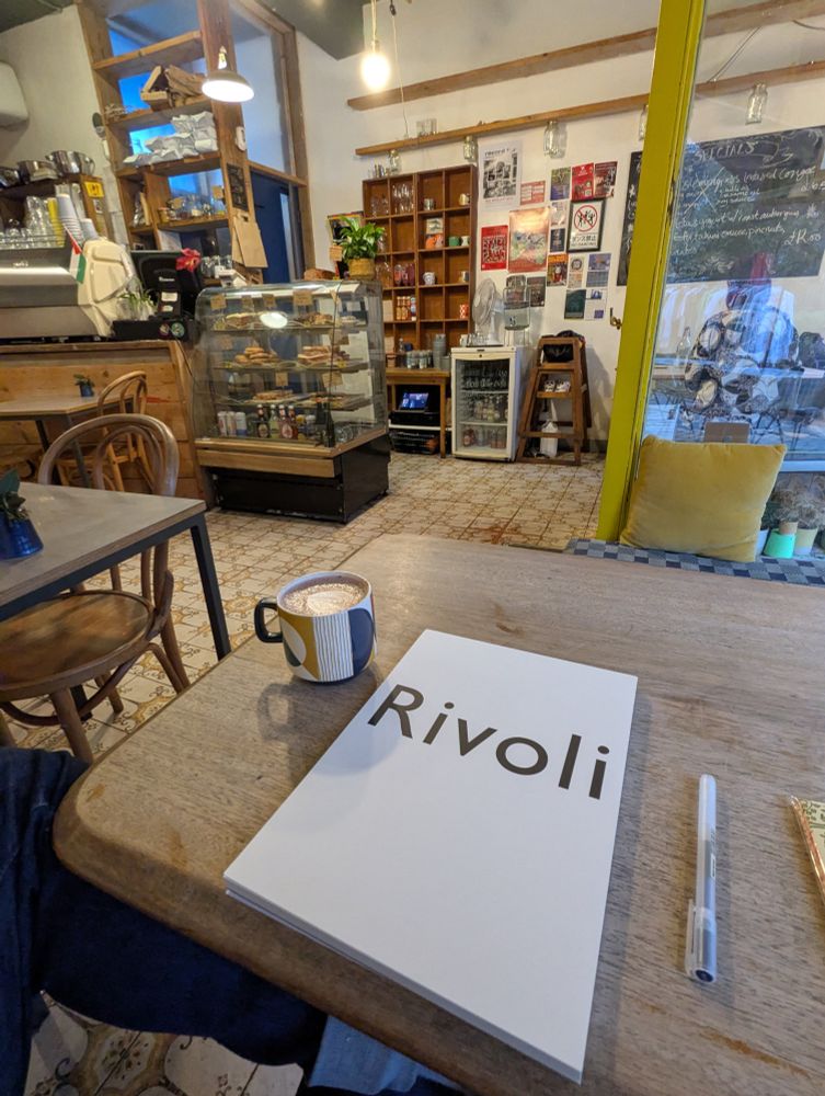 Interior view of Curious Yellow Kafe showing the full space with its glass display case of baked goods, espresso machine, wooden shelving units, and chalkboard menu on the wall. Rivoli letter writing paper and a hot chocolate in a geometric patterned mug sit on the rustic wooden table in the foreground. The café features mismatched vintage furniture, patterned tile flooring, exposed ceiling beams, and that distinctive bright yellow door frame. The eclectic, artistic vibe is enhanced by posters, plants, and warm lighting creating an inviting creative workspace.