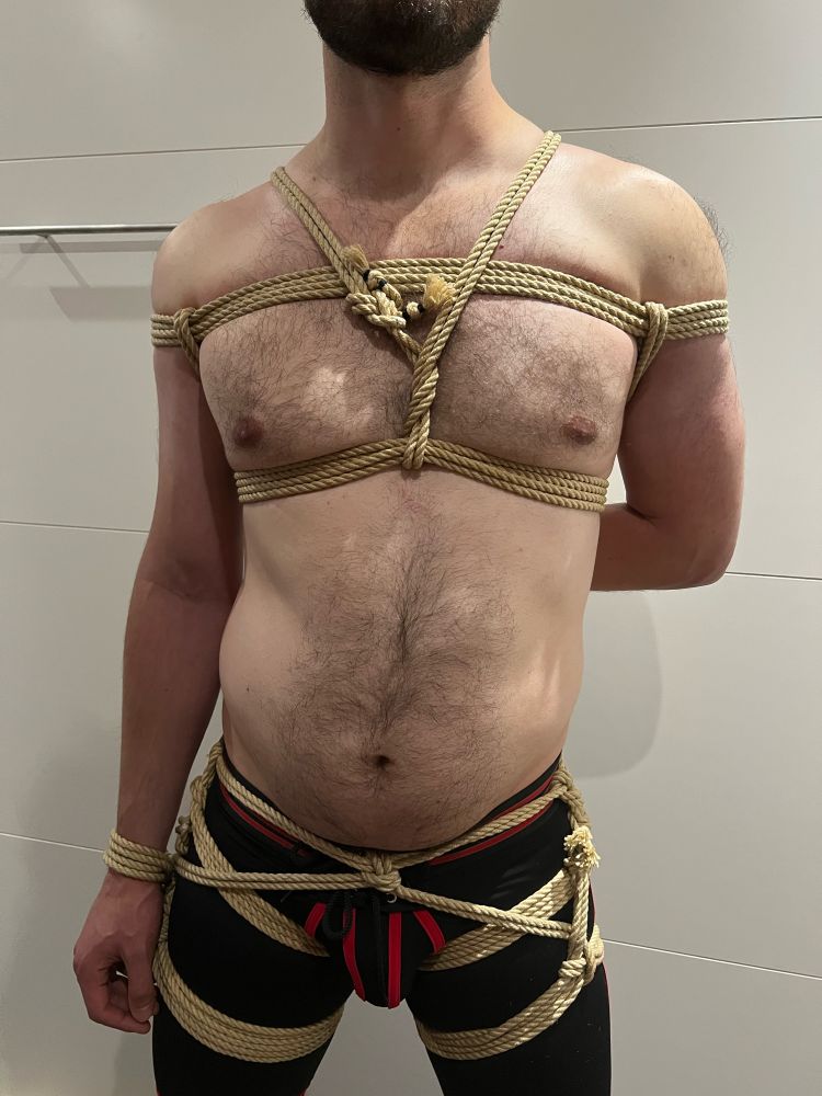A shirtless man stands against a plain background, bound in an intricate rope harness. The tan ropes wrap tightly around his chest, forming a symmetrical pattern across his torso and shoulders. His arms are secured behind his back, adding to the restrictive nature of the bondage. Additional ropes are tied around his waist, hips, and thighs, with a decorative knot arrangement over black shorts with red accents. The setup emphasizes both structure and restraint. The lighting is bright, and the setting appears minimal, with a smooth wall in the background.