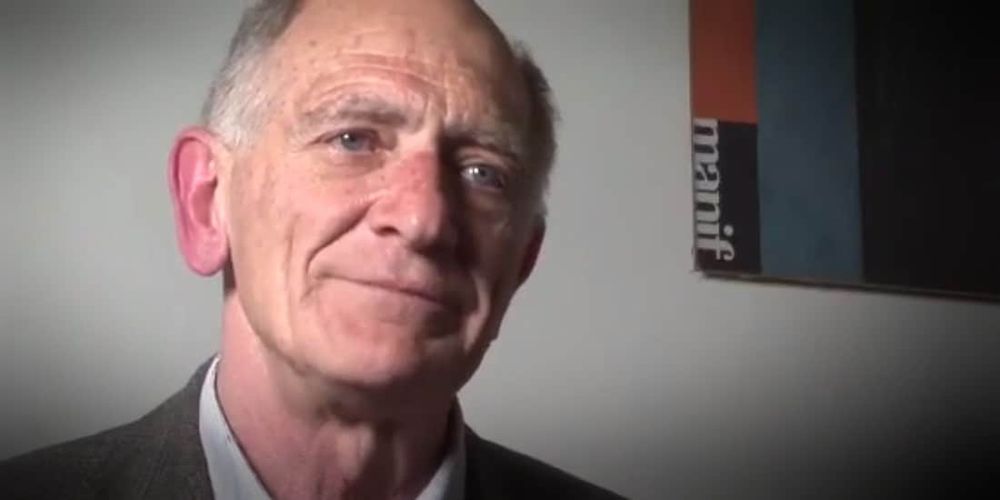 Un hombre mayor, sonriente, con poco pelo y sin barba. En el fondo, en un cuadro se lee "manif".