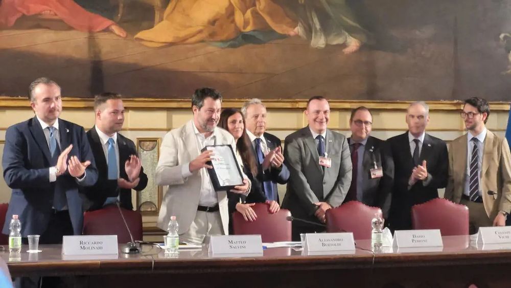 Matteo Salvini en traje blanco en una mesa rodeado de siete hombres y una mujer muestra una placa mientras sus compañeros le aplauden. Sonríe.