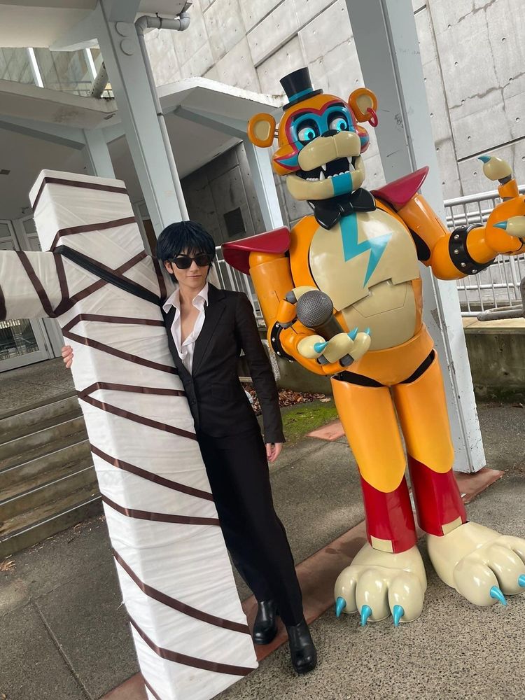 Photo of two costumers. One of them is dressed in a suit, portraying Wolfwood from Trigun, holding a large cross wrapped in fabric. The other is wearing a large orange robot bear costume, and he is portraying Glamrock Freddy from FNAF: Security Breach.