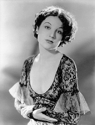 A black and white publicity photo of a young, beautiful, brunette woman. She is dressed in a scooped necked, lace dress, in a 1930's style.