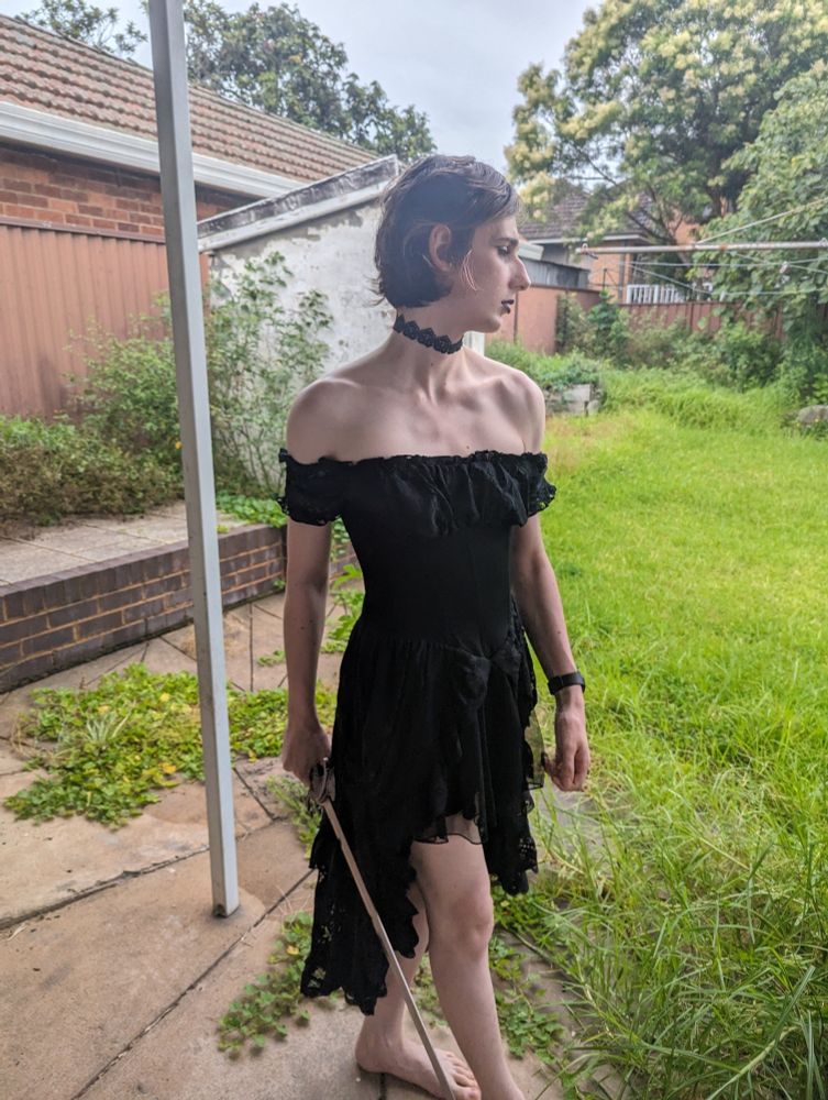 me in a black dress with exposed collarbones, wearing a black choker, looking to the side and holding a sword against a grassy backdrop