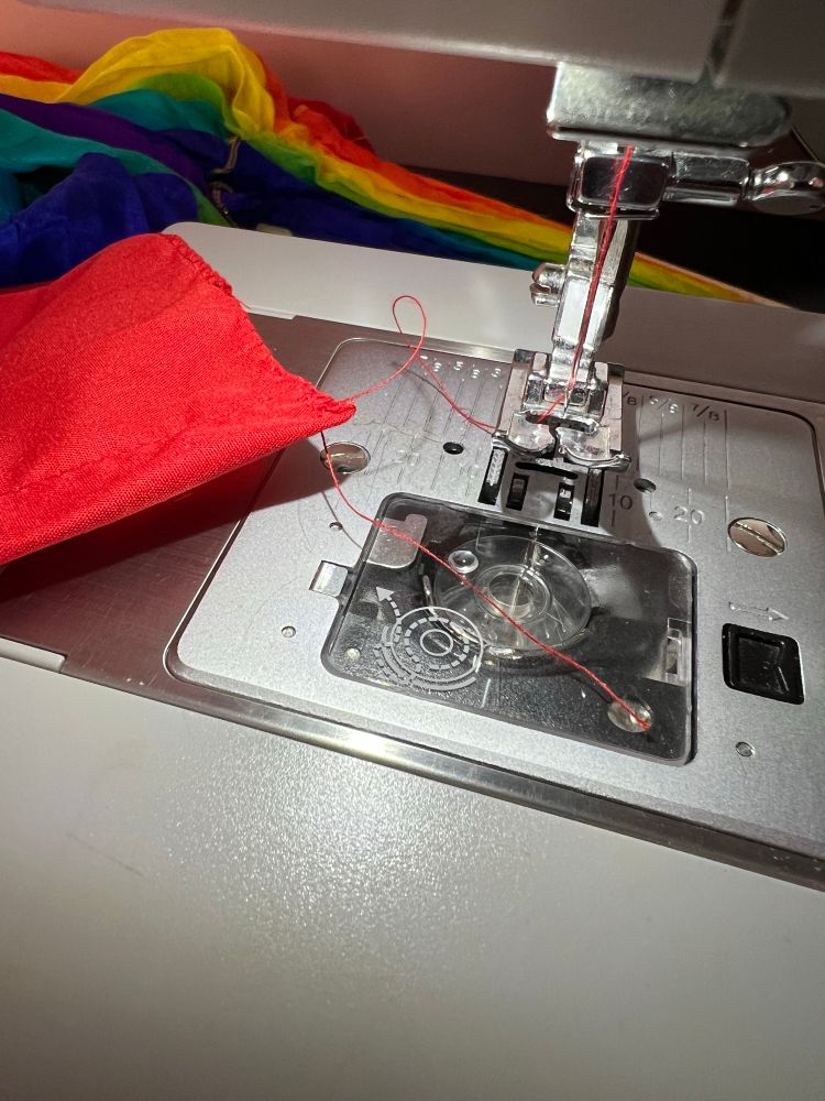 Close up of the foot on a sewing machine. An empty bobbin is visible through a transparent plate. Some red fabric is still on the needle thread and about 5cm of bobbin thread lies loose.