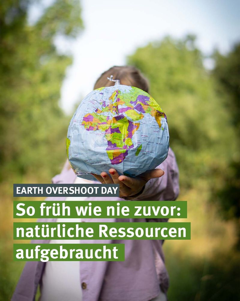 Eine Person hält eine zerknitterte, aufblasbare Weltkugel in die Kamera, sodass sie den eigenen Kopf vollständig verdeckt. Die Weltkugel zeigt Afrika, Europa und Teile Asiens. Im Hintergrund ist grüne Natur unscharf zu erkennen. Auf dem Bild steht in weißen und grünen Textfeldern: „EARTH OVERSHOOT DAY – So früh wie nie zuvor: natürliche Ressourcen aufgebraucht“.