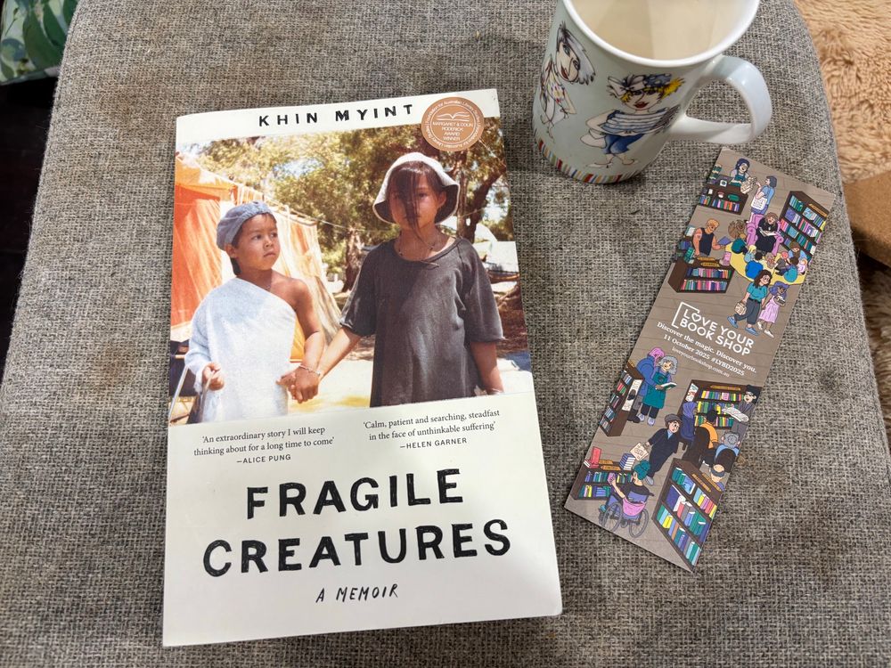 Book: Fragile Creatures by Khin Myint. Photographed on a grey fabric stool that also has a coffee mug and a “love your bookshop” bookmark 