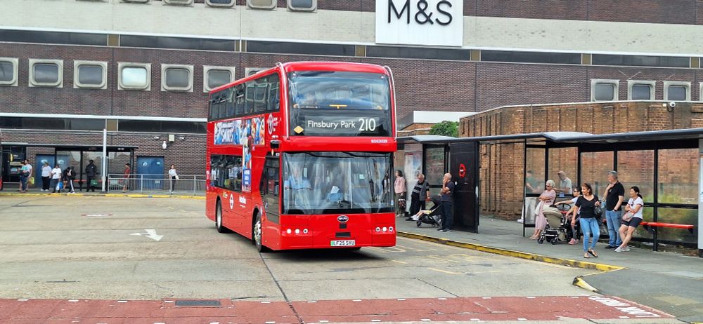 A red double decker bus with Shopping Centre building behind. People waiting at bus stops.