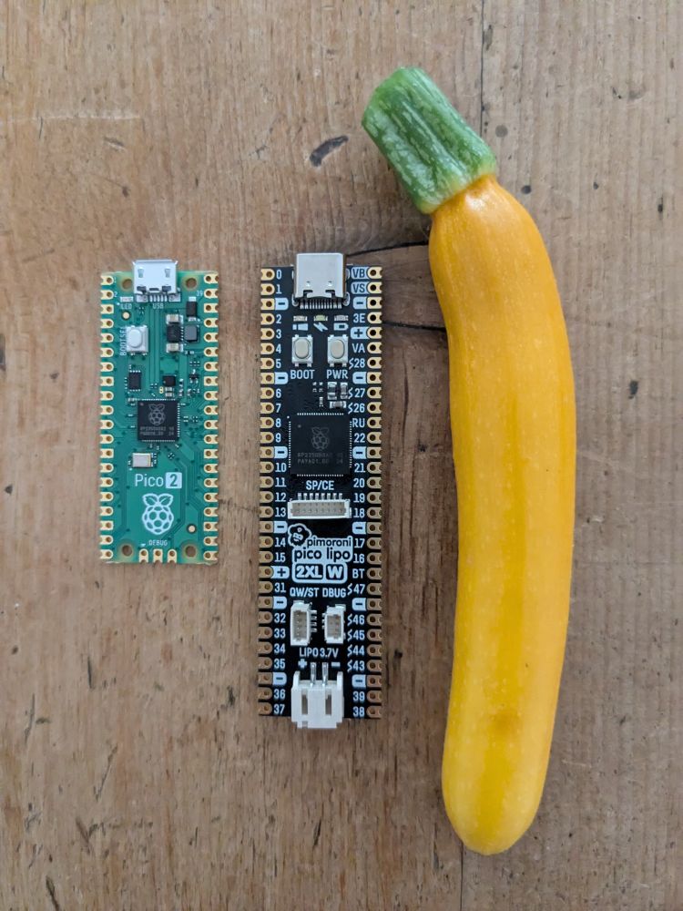 On a wooden tabletop are laid out: a Pimoroni Pico LiPo (2 XL W) in the centre of the frame, a Raspberry Pi Pico 2 to its left, and a small, yellow, home-grown courgette (zucchini) to its right. Pimoroni's Pico (a microcontroller development board) is about one and a half times the length of Raspberry Pi's Pico, and both Picos are smaller than the courgette.