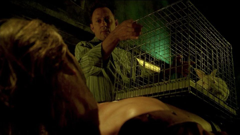 A man holds a caged rabbit atop the bare torso of another man