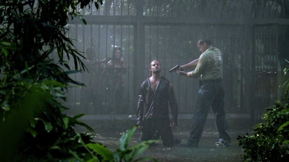 A man kneels in the rain while another man points a gun at him, and a woman held at gunpoint watches from a cage