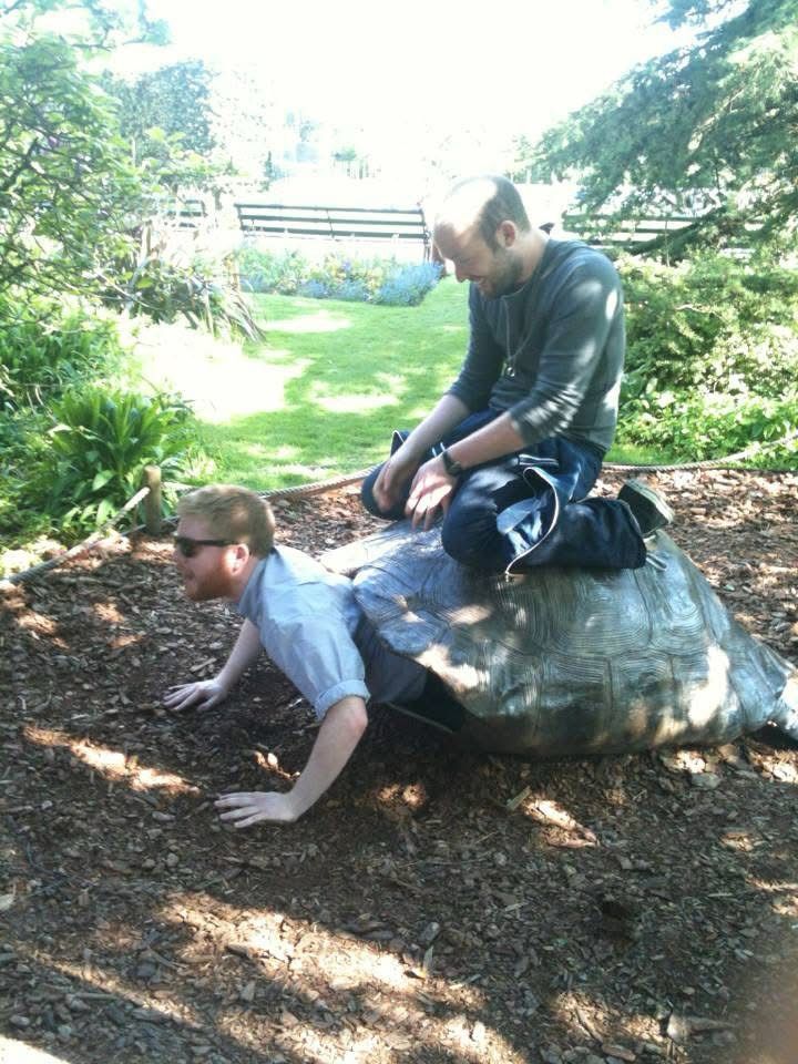 Alan, smiling coyly astride a model giant tortoise shell, from who’s neck hole Jon emerges wearing sunglasses.