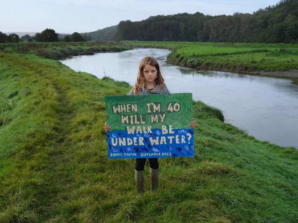 A ten-year-old standing next to a river in a North Devon community near a tidal zone. She is standing on land which is projected to be affected by regular climate change-related flooding by 2050. She is holding a hand-made sign reading "When I'm 40, will my walk be under water?"
