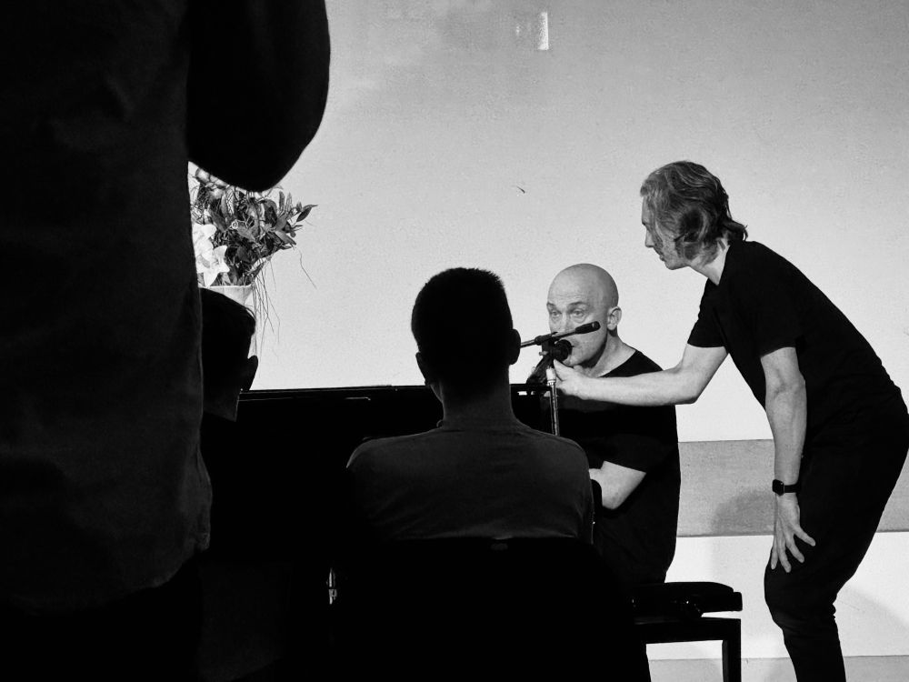 Chris Thorpe playing piano and singing while a technician holds a microphone for him 