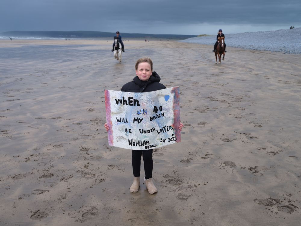 A ten-year-old on a beach under leaden skies near a pebble ridge, with two horses being ridden towards her in the background. She is holding a sign reading "when I'm 40 will my beach be underwater?"