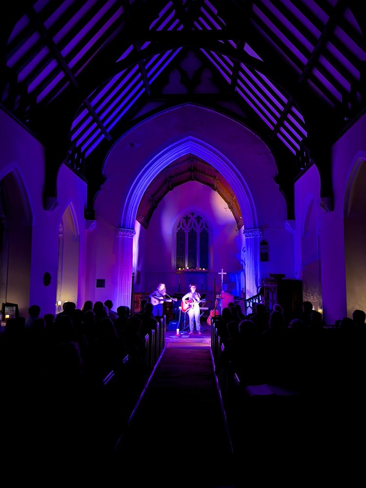 Two accomplished female musicians playing an evening acoustic gig at the front of a rural parish church, lit by candles in the window alcoves and a few floor lights. The audience fills the pews.