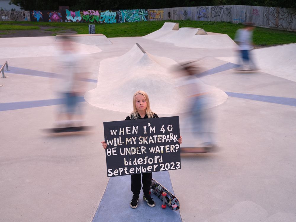 A ten-year-old in a skate park near a river. The skateboarders behind him are blurred due to movement. He is holdin a sign reading "When I'm 40 will my skatepark be underwater?"