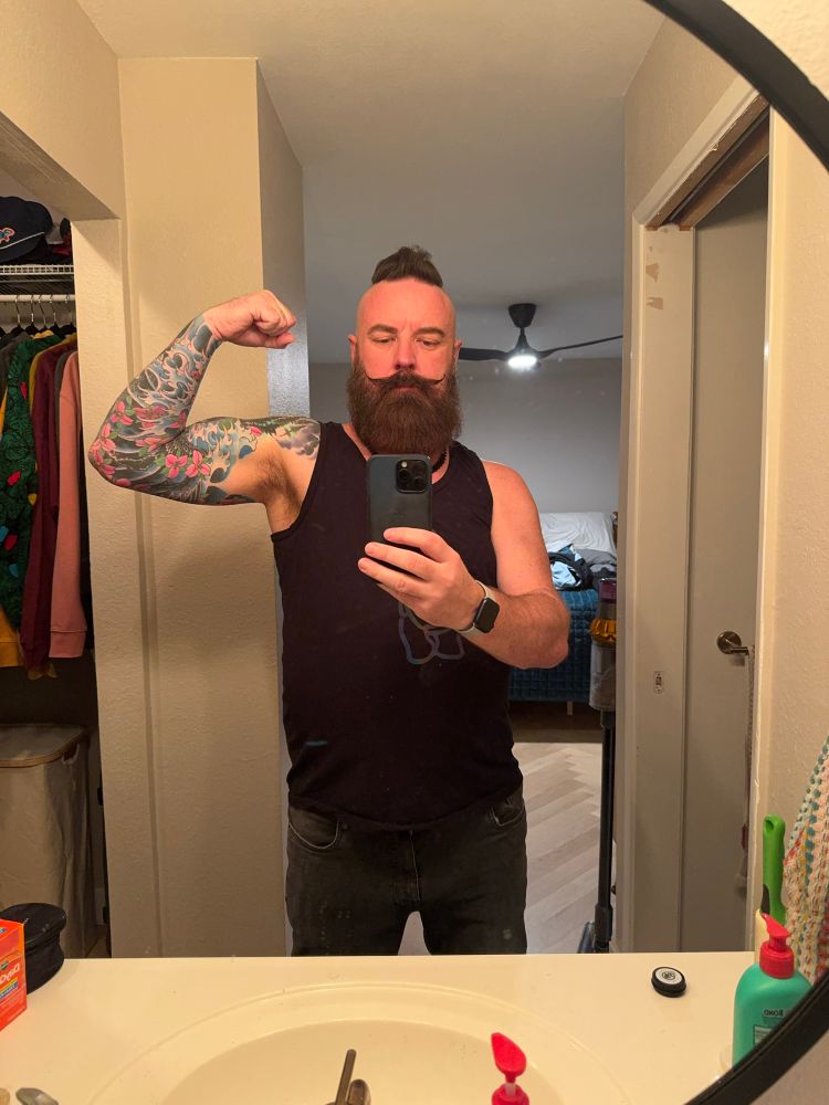 White male with sleeve tattoo and large beard flexes a bicep in a mirror. 