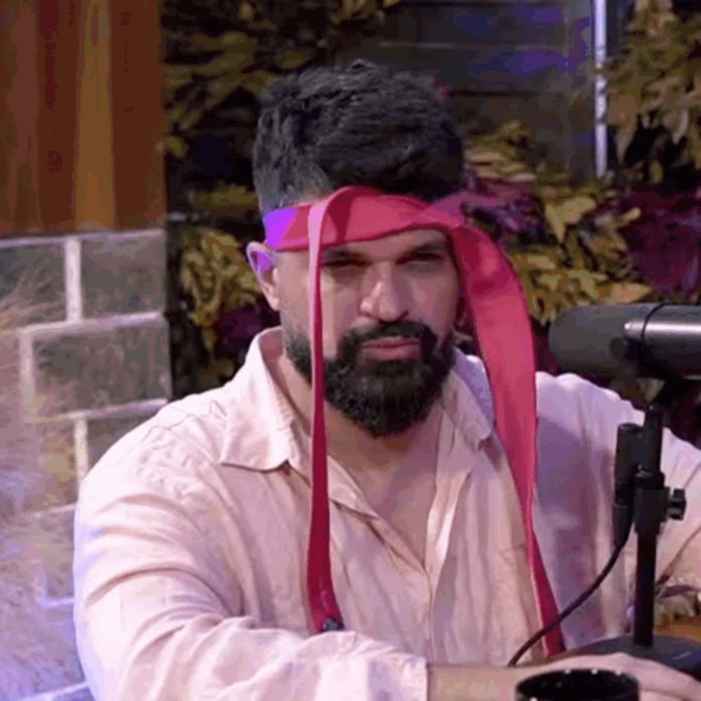 a man with a beard and a red headband is sitting in front of a microphone and looking at the camera .