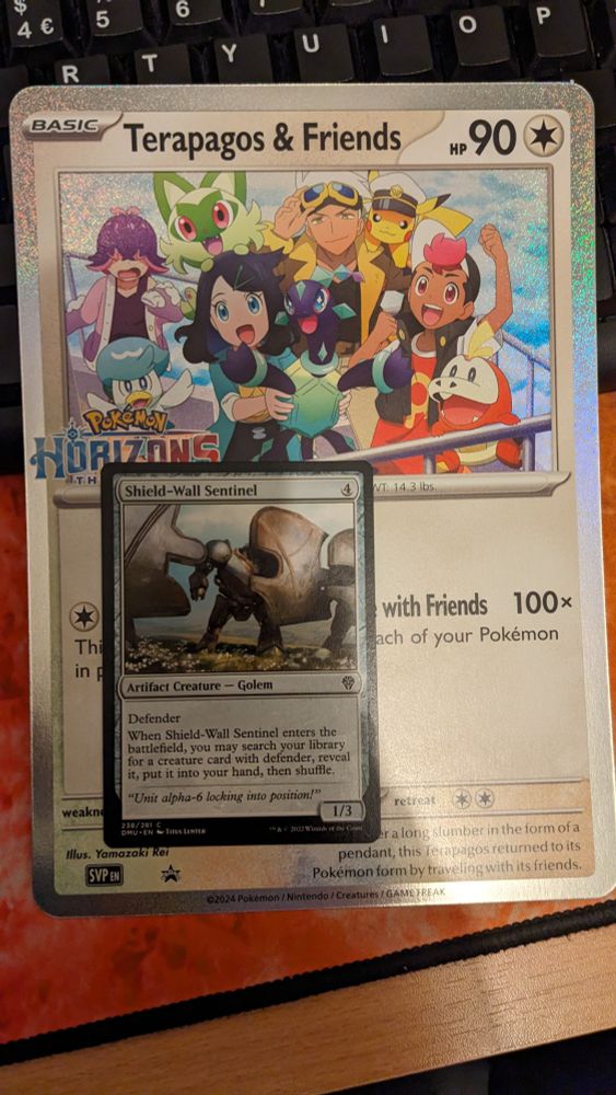 Same promotional Pokémon card from before "Terapagos & Friends", but with the Magic The Gathering card "Shield-Wall Sentinel" placed over it to compare scale. The jumbo Pokémon promo  is at least 2.5x larger than the Magic card.