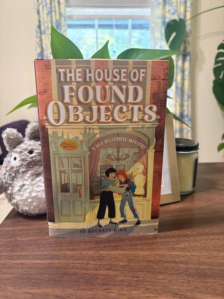 Copy of THE HOUSE OF FOUND OBJECTS standing on wooden desk with a stuffy of Totoro, a plant, and a window in the background.